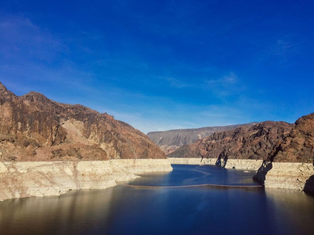 Photo of Lake Mead, showing deep water rings on the rocky banks caused by falling water levels, under a deep blue sky.